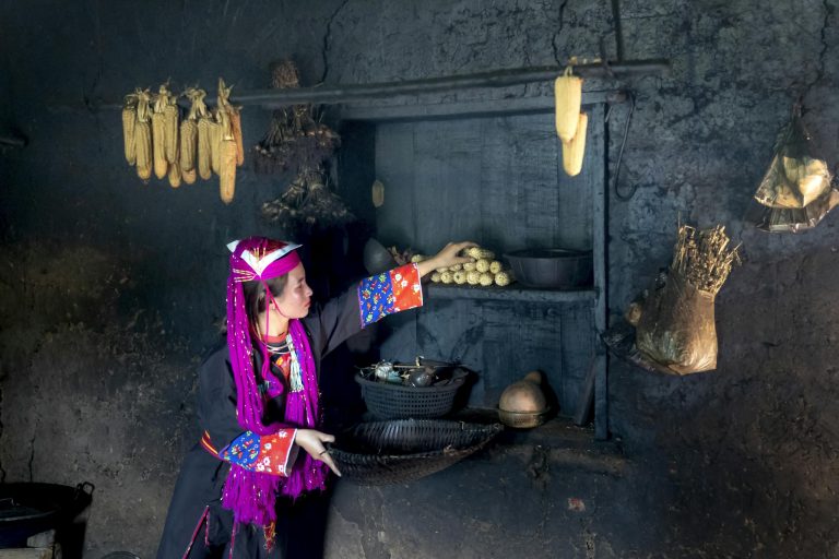 Woman in traditional attire reaching for corn in a rustic storage room filled with food items.