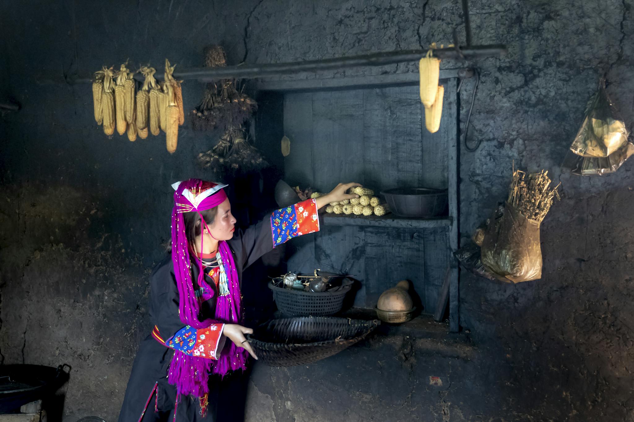 Woman in traditional attire reaching for corn in a rustic storage room filled with food items.